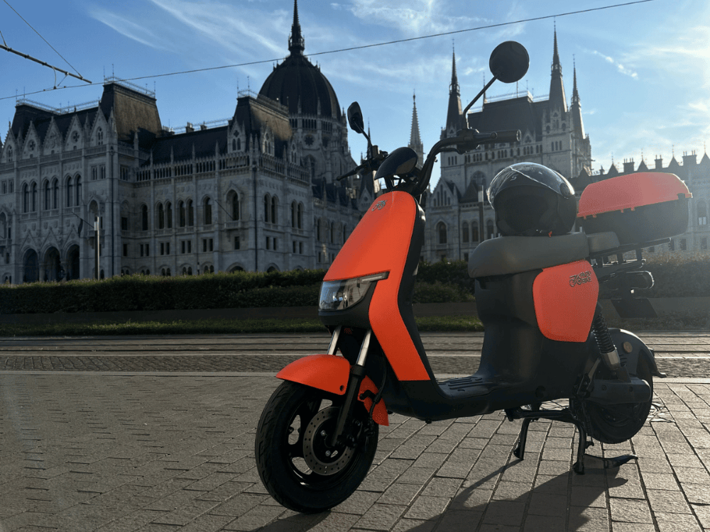Narancssárga elektromos robogó a budapesti Parlament előtt, modern dizájnnal és hátsó tárolódobozzal, park mód funkcióval a biztonság és energiahatékonyság érdekében, készpénzes vagy részletfizetéses vásárlásra elérhető robogó kereskedésünkben.