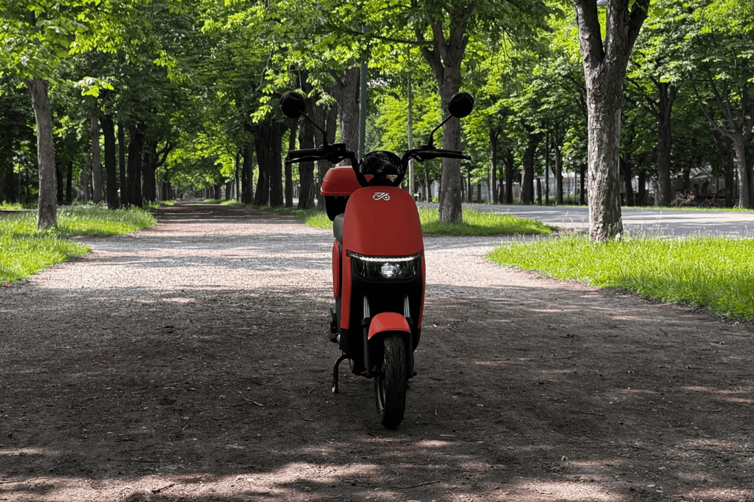  Narancssárga elektromos robogó eladó vagy bérelhető, erős akkumulátorral, ideális városi közlekedéshez, parkolva egy nyári budapesti parkban, földes úton, fák között